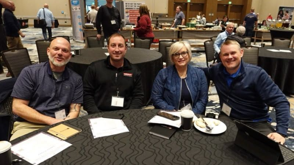 Four people smiling at a round table in a restaurant, with plates, drinks, and a conference lanyard visible