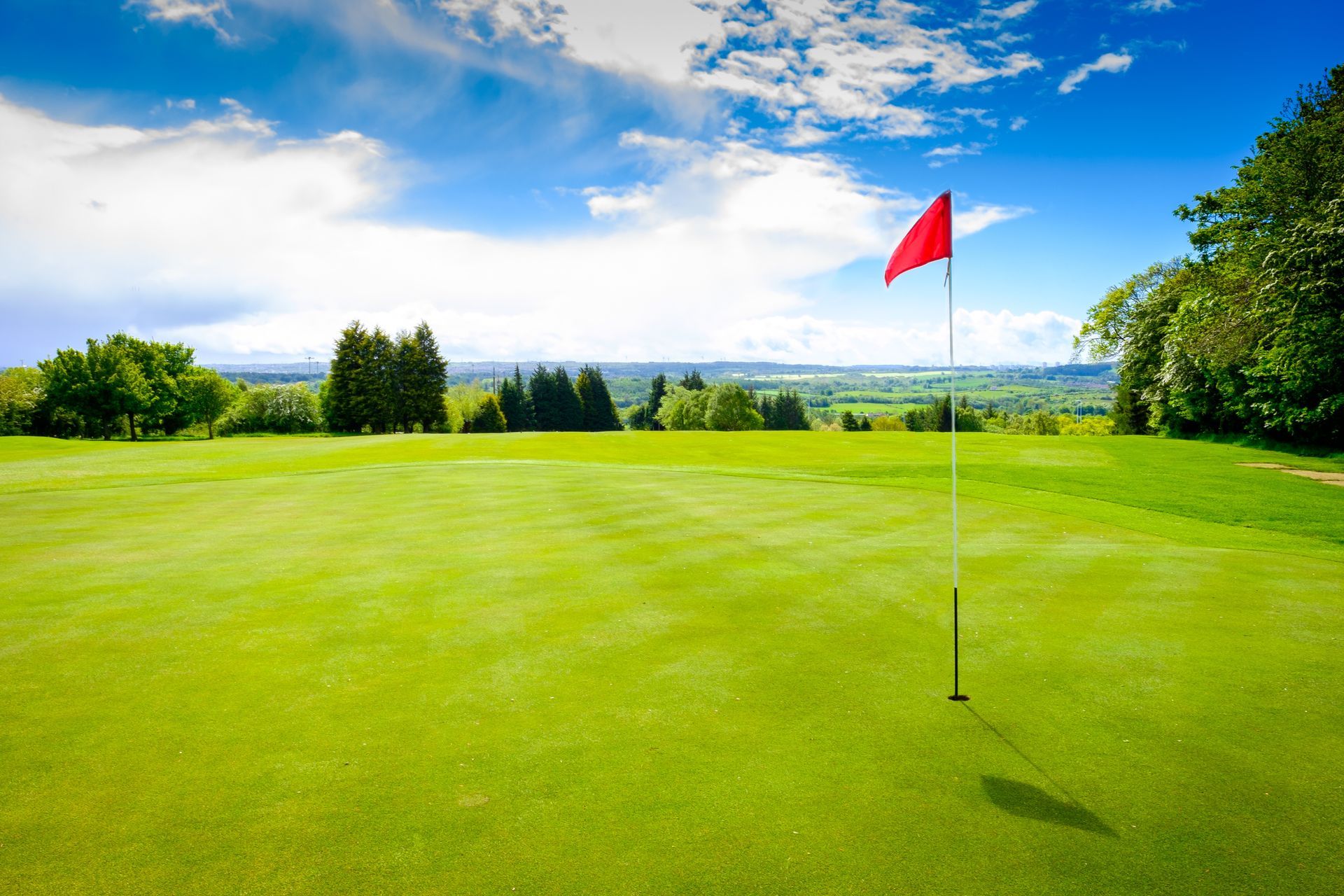 Golf green with a red flag under a bright blue sky with clouds