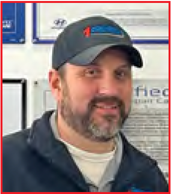 Smiling man in a dark jacket and cap standing indoors against a wall with framed certificates