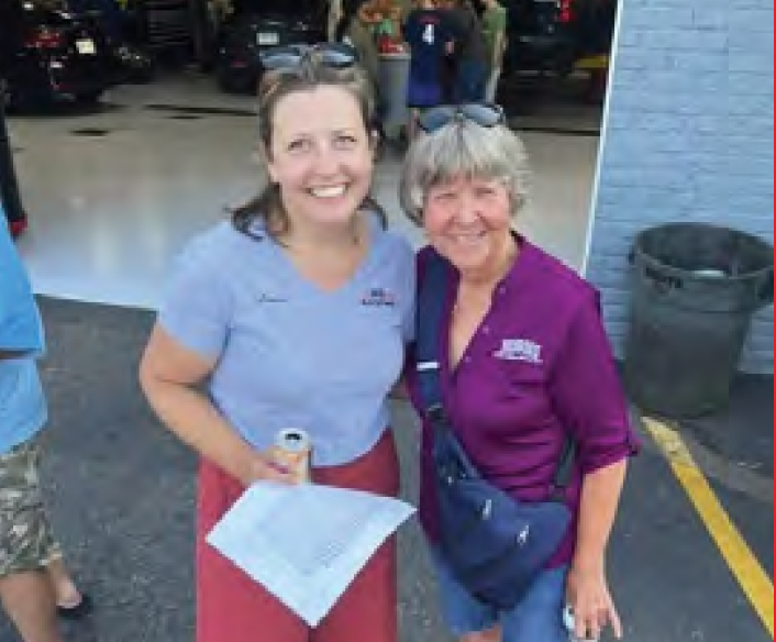Two smiling people stand in a garage, one holding a drink and paper, with cars and a trash can behind them.