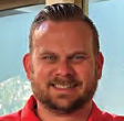 Smiling man with short hair and beard wearing a red shirt, outdoors with a blurred green background