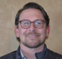 Smiling man with glasses and dark jacket against a tan background