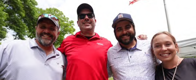 Four smiling people posing outdoors, two wearing caps and one in a red shirt.