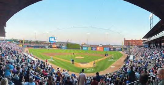 Crowded baseball stadium at dusk, with fans filling the stands around the field and scoreboard.