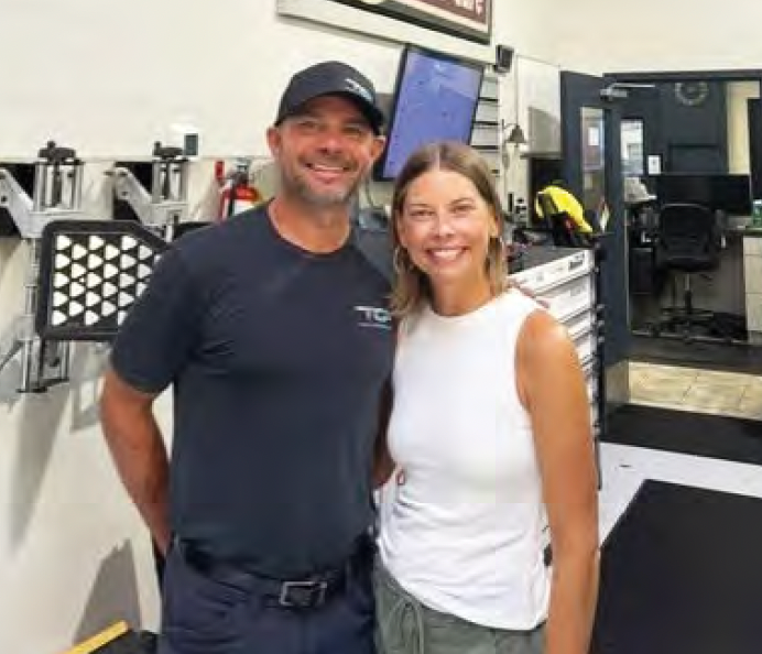 Two smiling people standing together in a gym, with workout equipment and mirrors behind them.