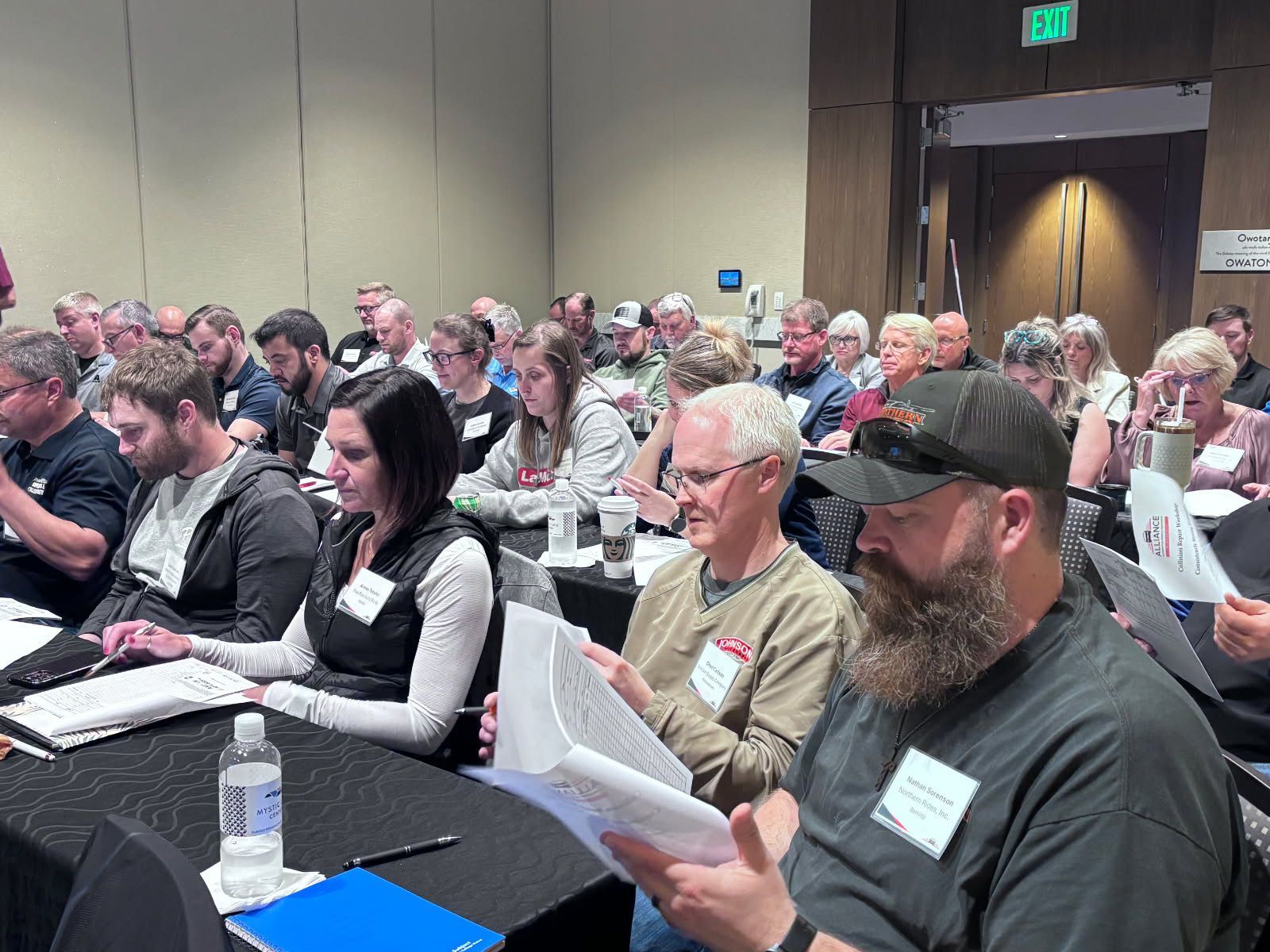 Audience in a conference room; people seated at tables, reading materials, some with name tags.