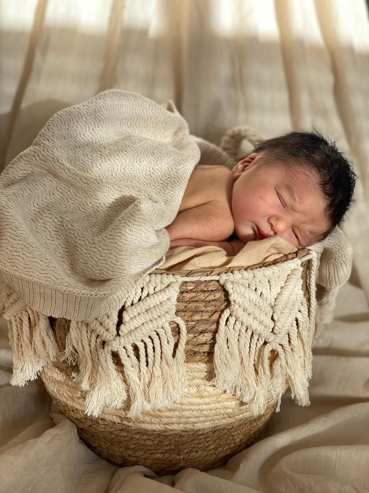 Een slapende baby, gewikkeld in een crèmekleurige deken, ligt in een gevlochten mand, met een zachtbeige achtergrond.