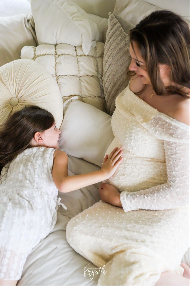 Vrouw en kind in witte jurken liggen op een bed en raken een zwangere buik aan.