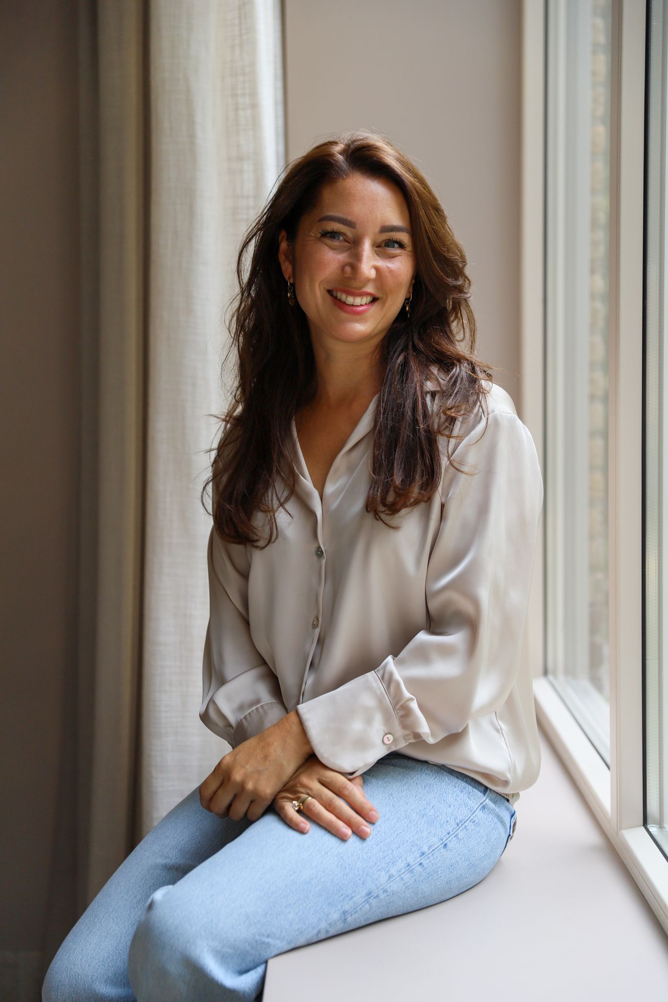 Vrouw in een lichte blouse en jeans zit op de vensterbank en lacht in het natuurlijke licht.
