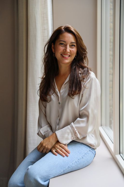 Vrouw in een lichte blouse en jeans zit op de vensterbank en lacht in het natuurlijke licht.