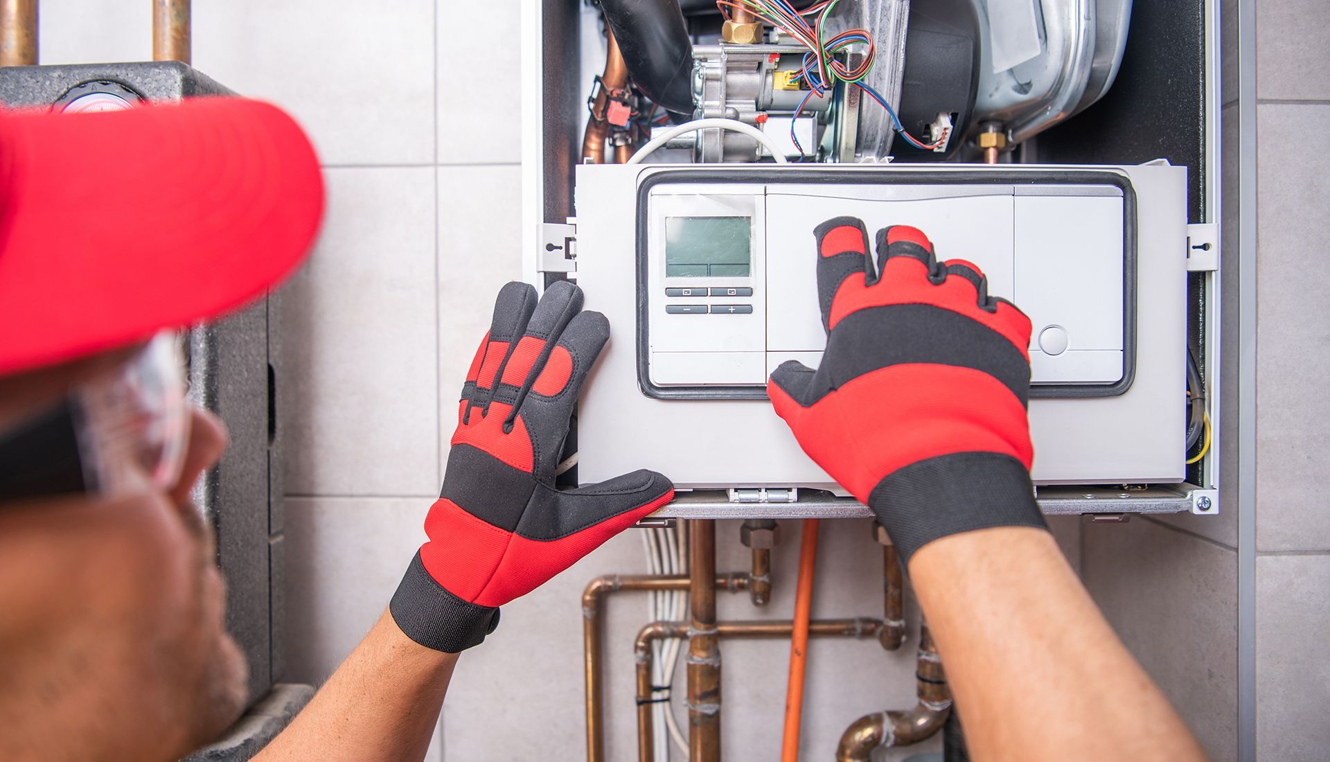 A technician in a red hat and work gloves inspects the control panel of a wall-mounted boiler.