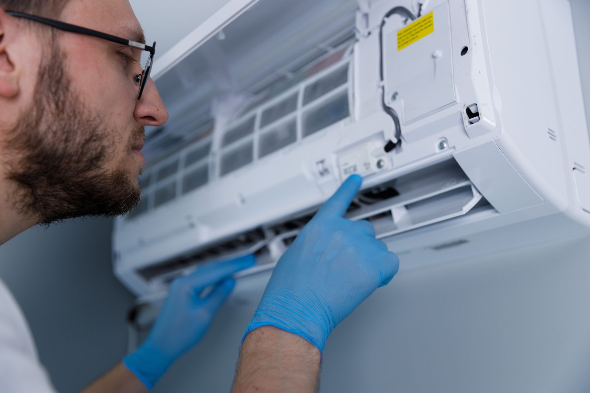 A technician wearing blue gloves cleans the interior components of a white wall-mounted air conditioner.