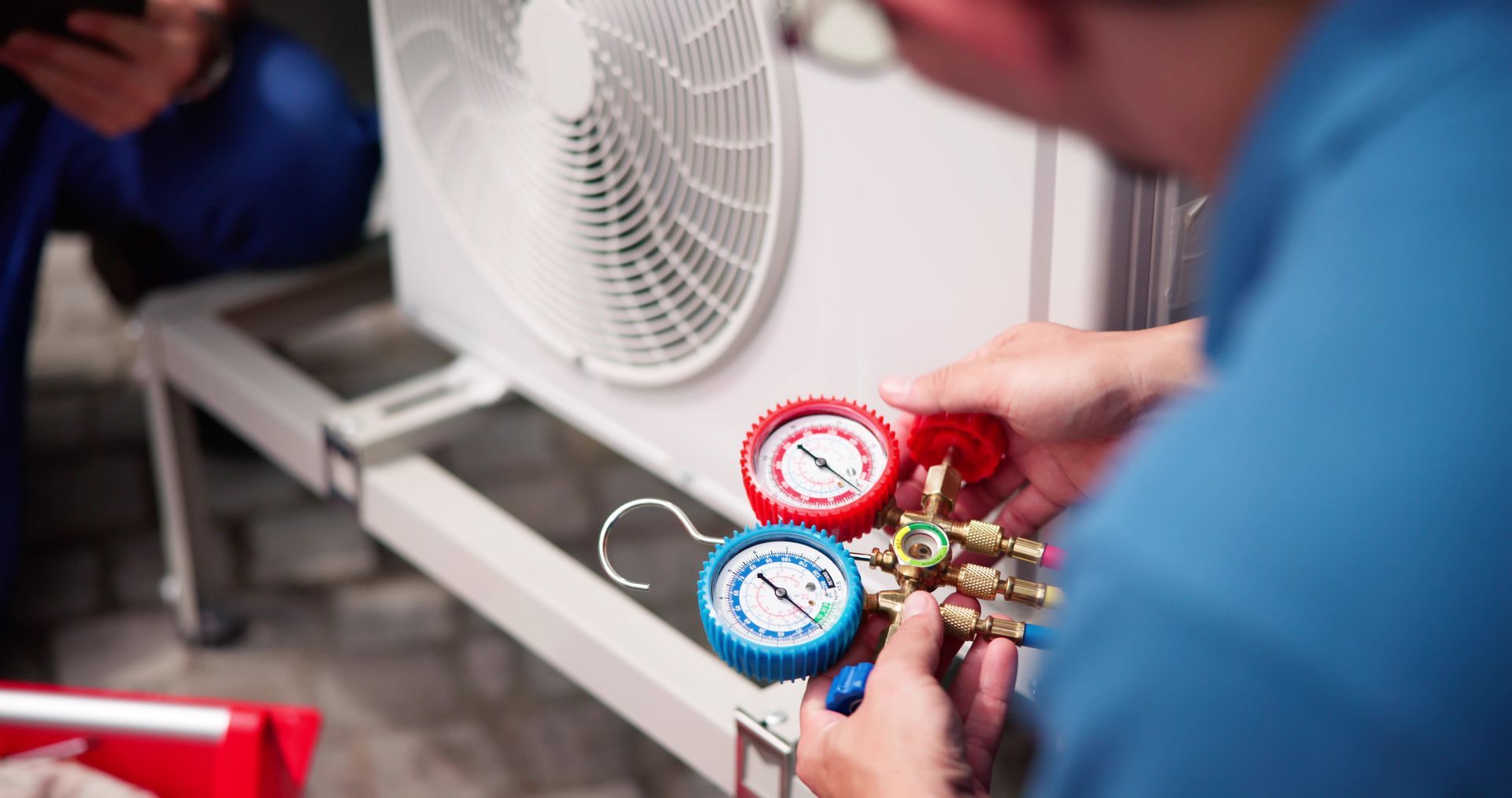 A person in a blue shirt uses a manifold gauge set to service an outdoor air conditioning unit.