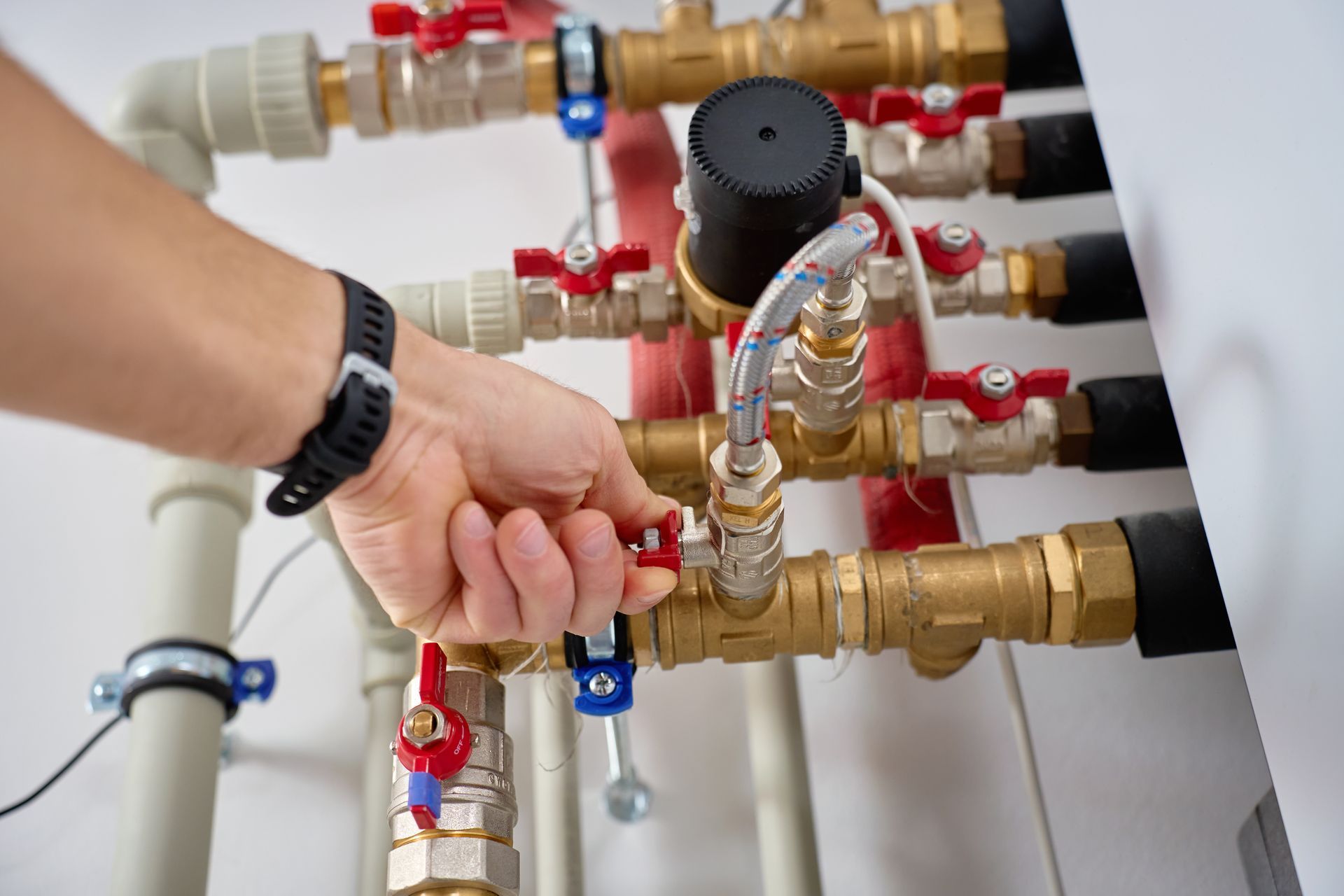 Hand adjusting a brass water manifold with red and blue valve handles on white pipes