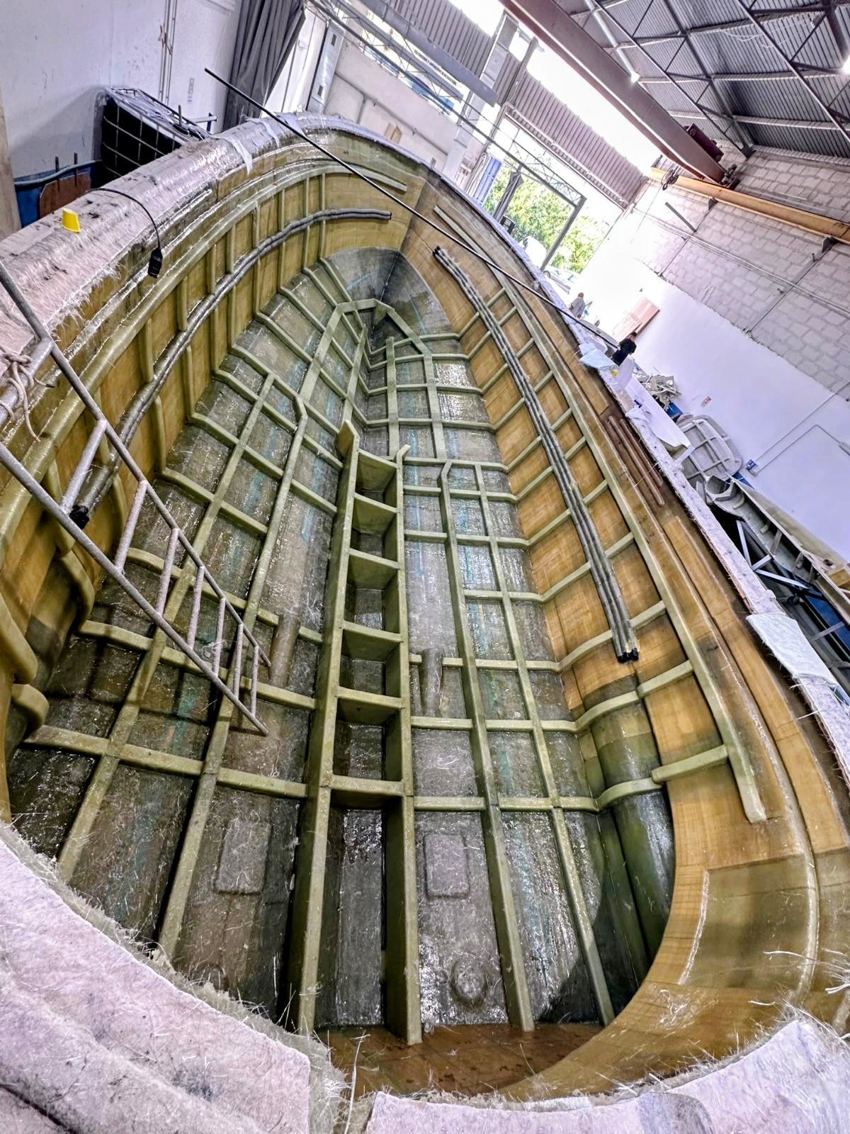 Lo scafo di un motoscafo in fibra di vetro bianca, non rifinito, con finiture del ponte in legno, è parcheggiato in un cantiere navale industriale all'aperto.