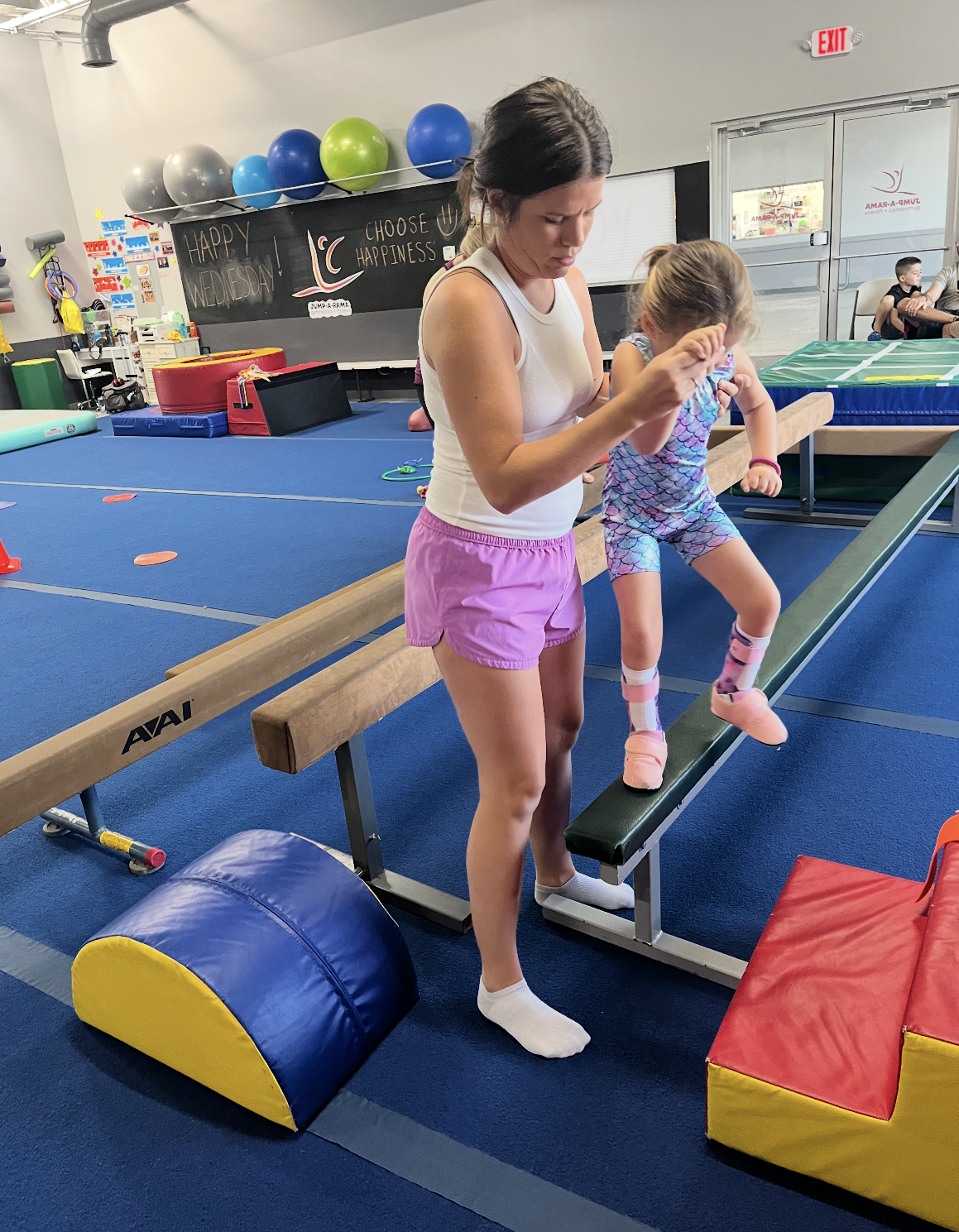 A woman is helping a little girl balance on a balance beam in a gym.