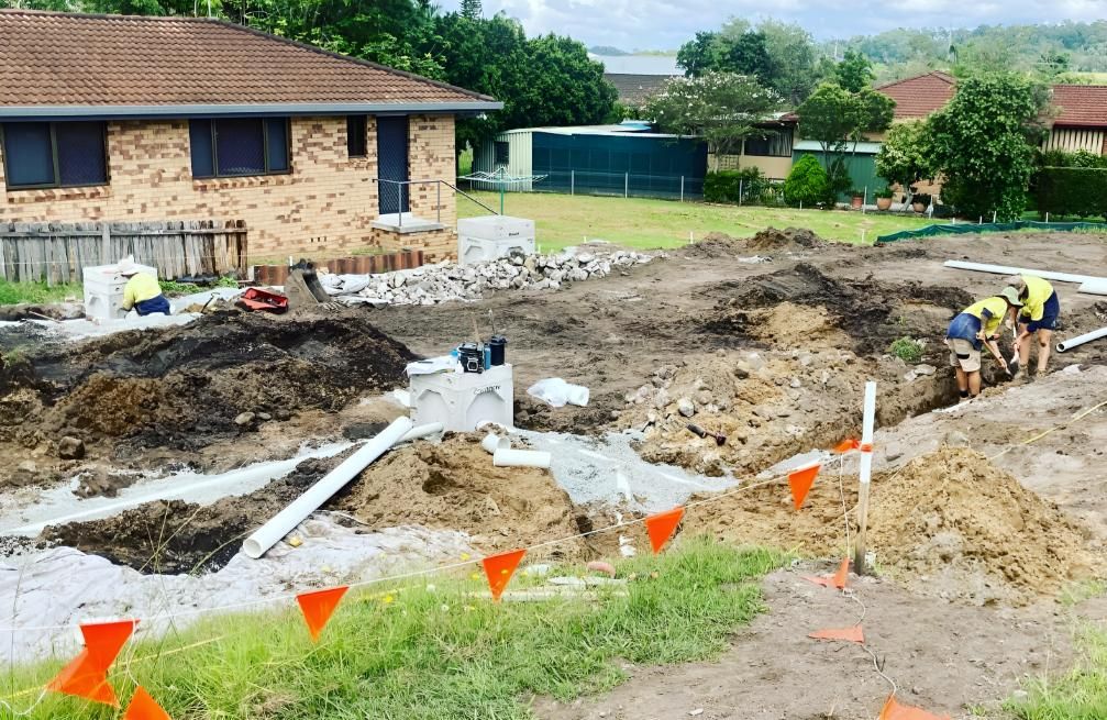Construction site: Workers digging trenches near a house, with pipes and dirt piles visible.
