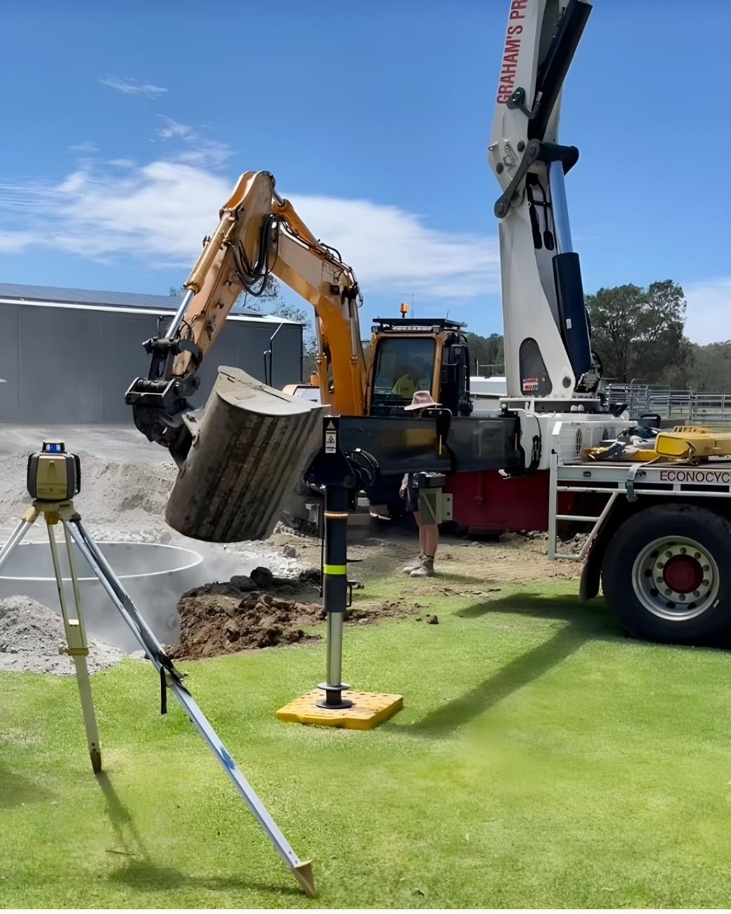 An Excavator is Digging a Hole in the Grass Next to a Truck — Shawline Plumbing and Drainage In Iluka, NSW