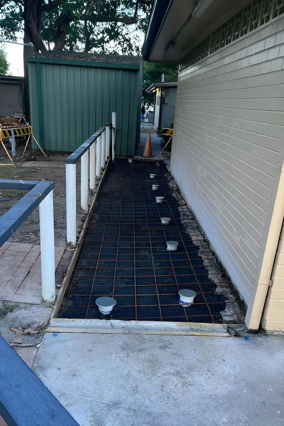 Construction site: concrete path framed with rebar and pipe fittings, next to building and railing.