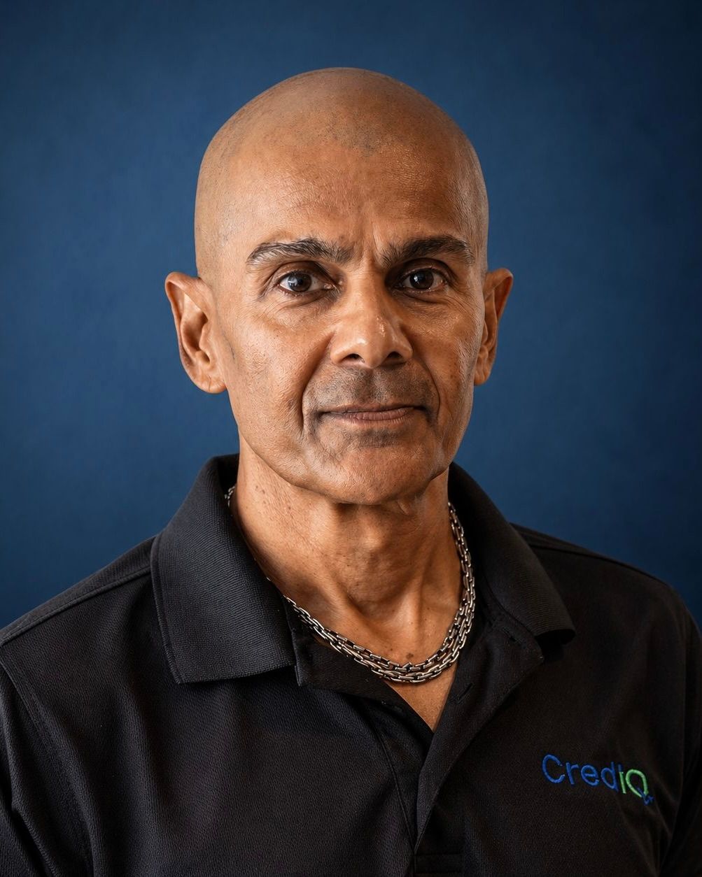 Razeem Ali wearing a dark polo shirt with a chain necklace, posed against a light, textured background.