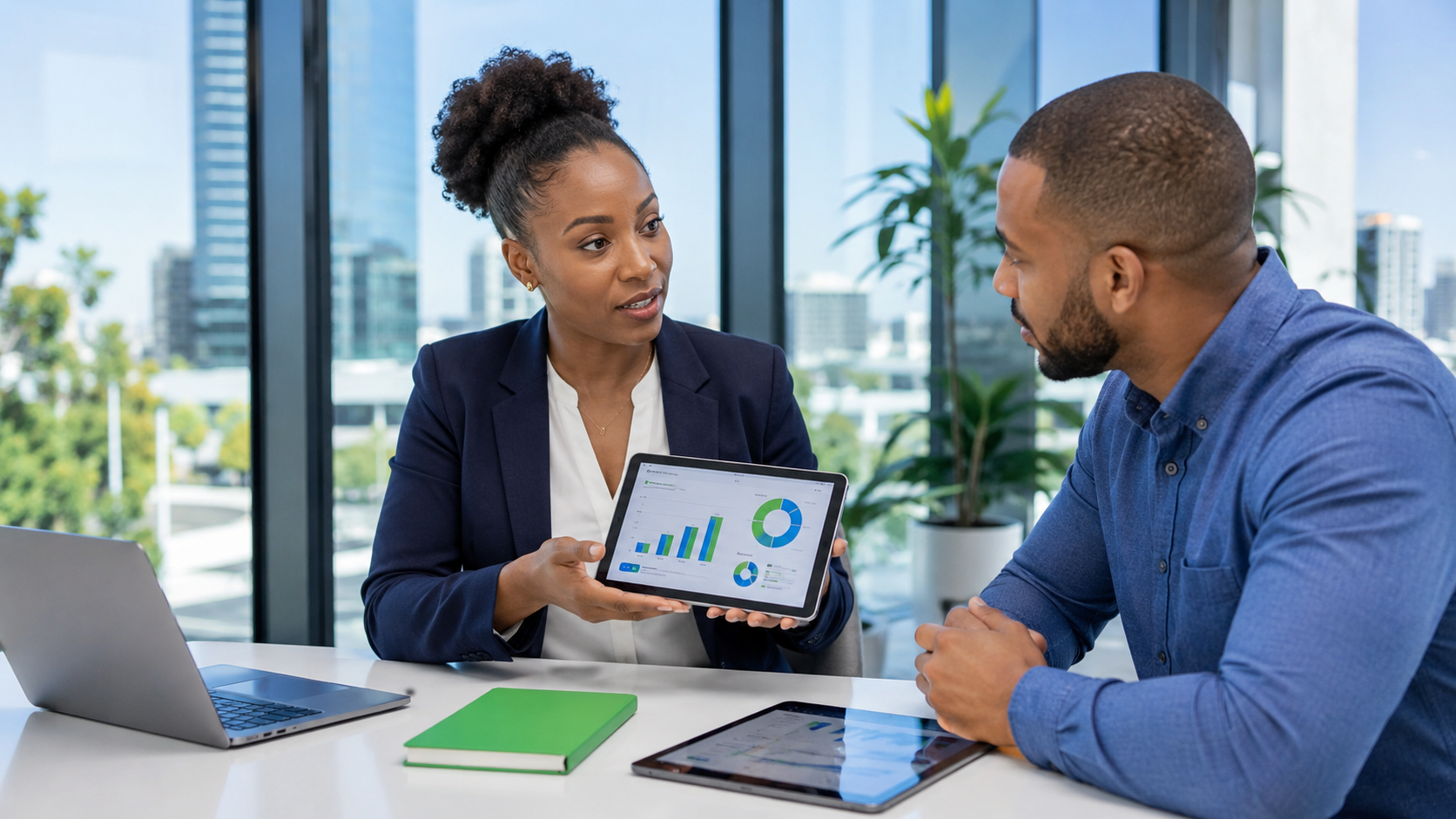 Two coworkers in a bright office reviewing a laptop and tablet with charts on the screen