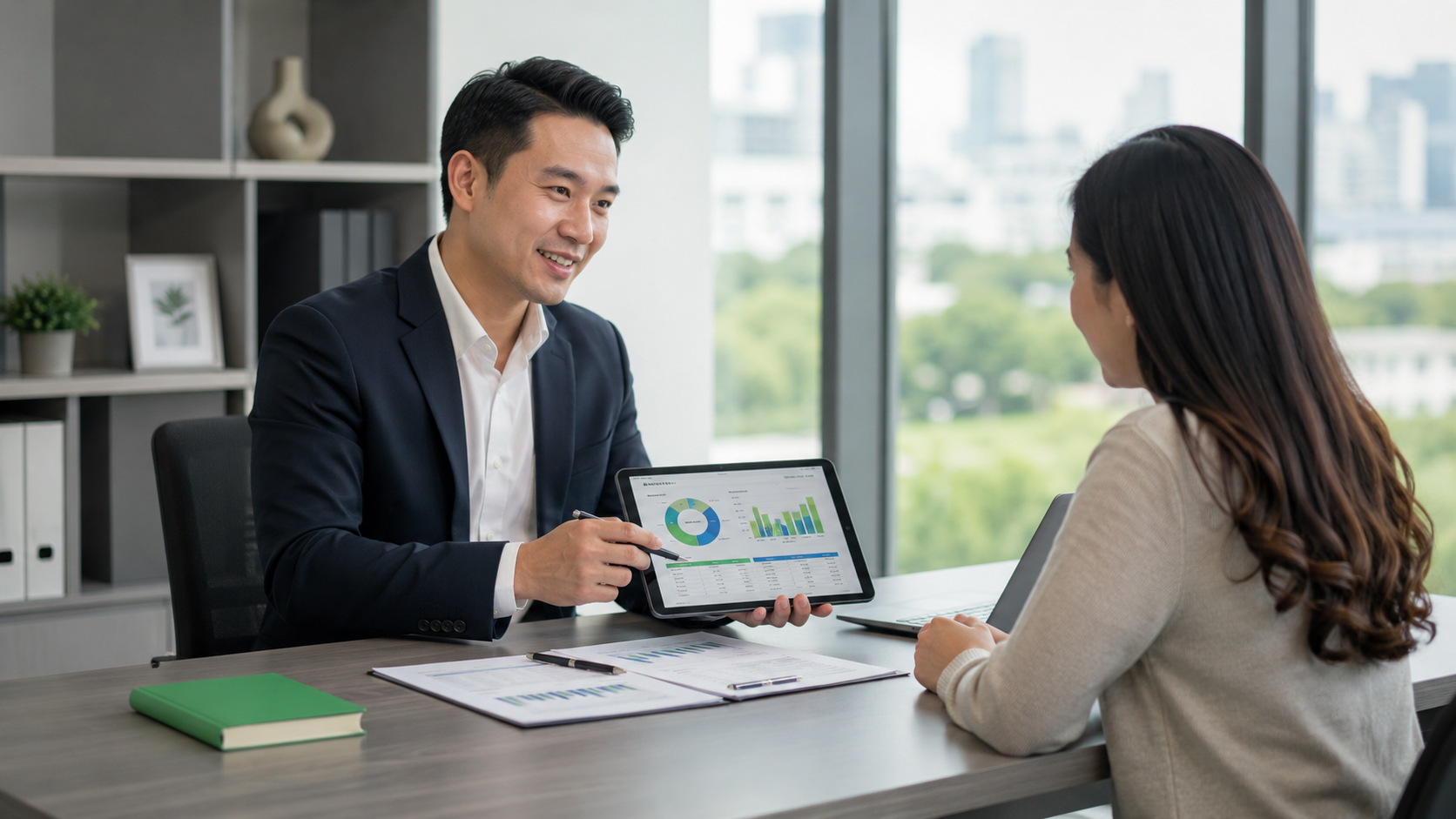 Two people discuss charts on a tablet across a desk in a bright office.