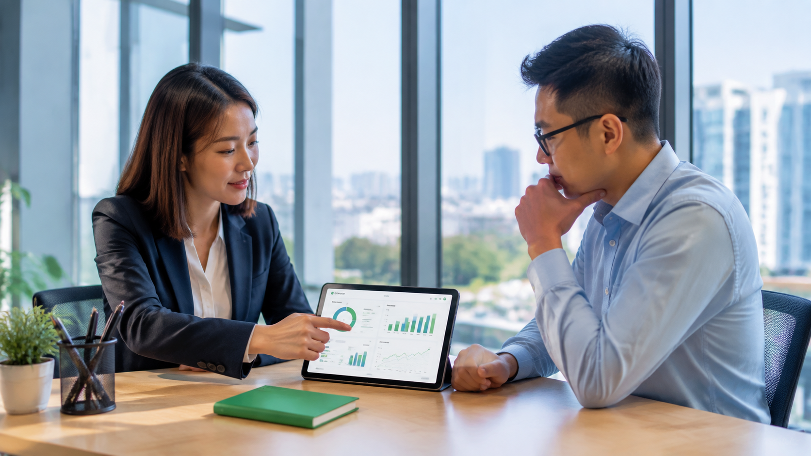 Two coworkers discussing a laptop chart in a bright office, with a notebook and coffee on the table.