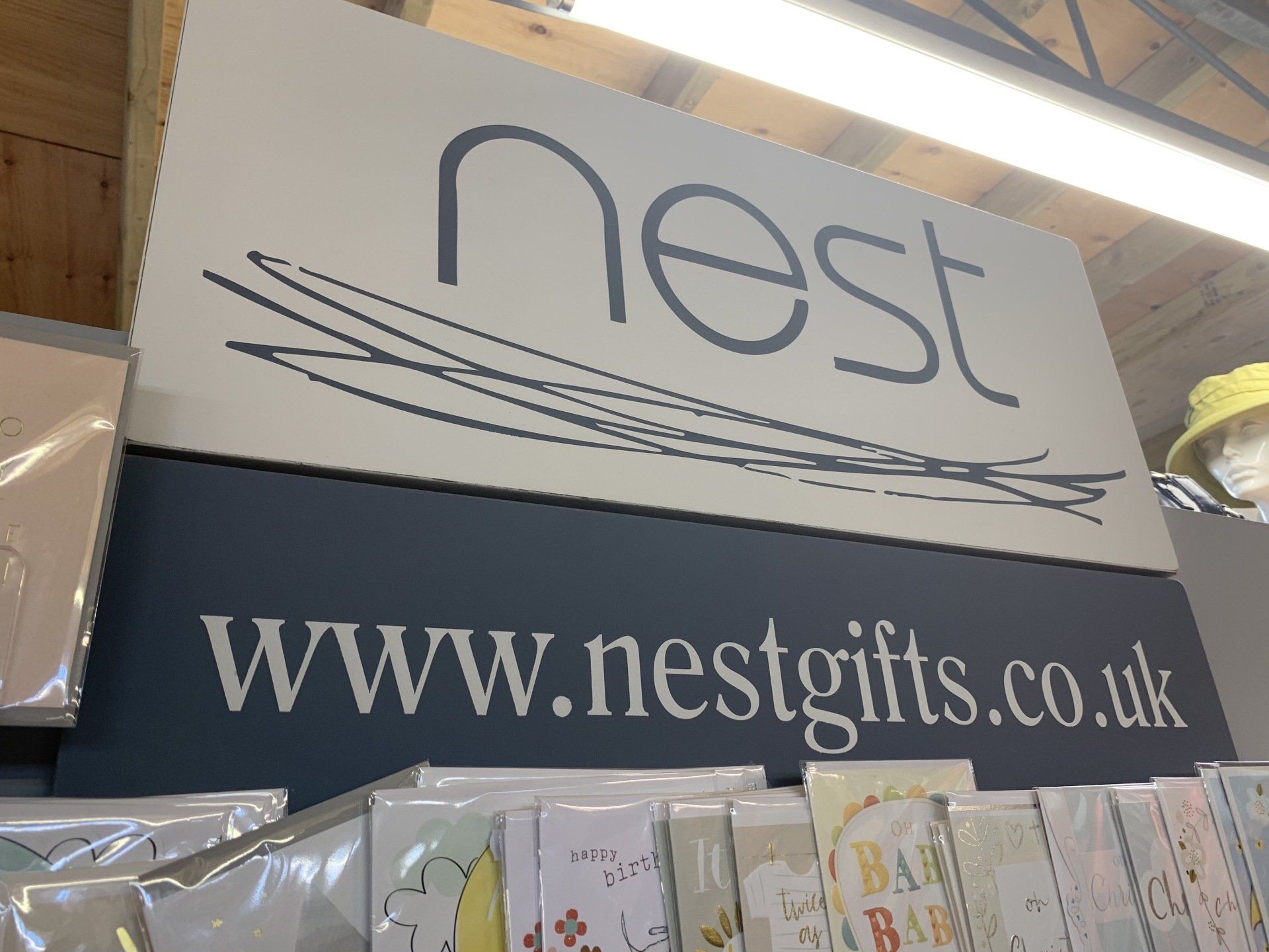 Sign for Nest Gifts store, with logo and website address, over greeting cards.