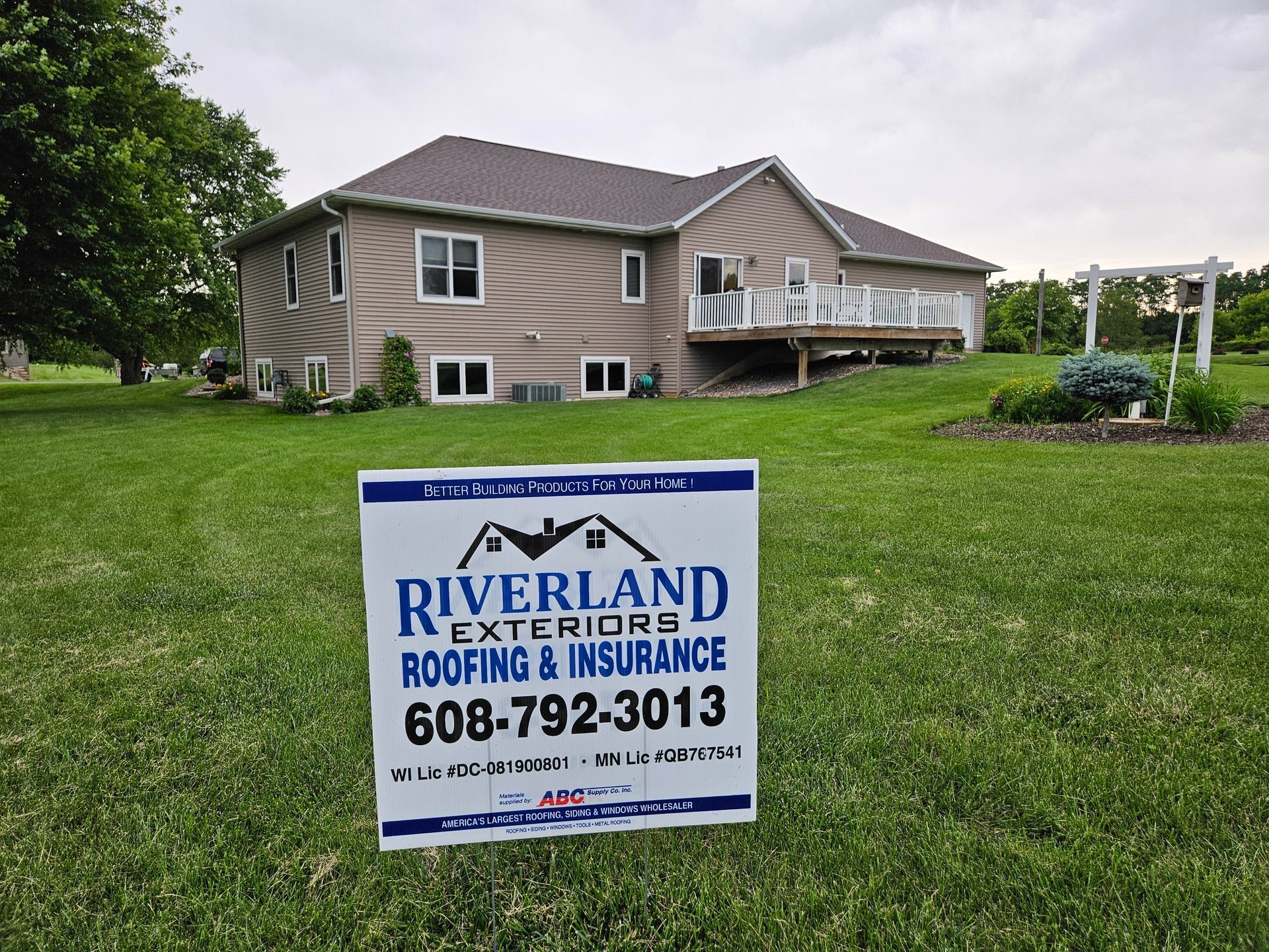 A sign for Riverland Exteriors in front of a tan two-story house with a brown roof and a white deck.