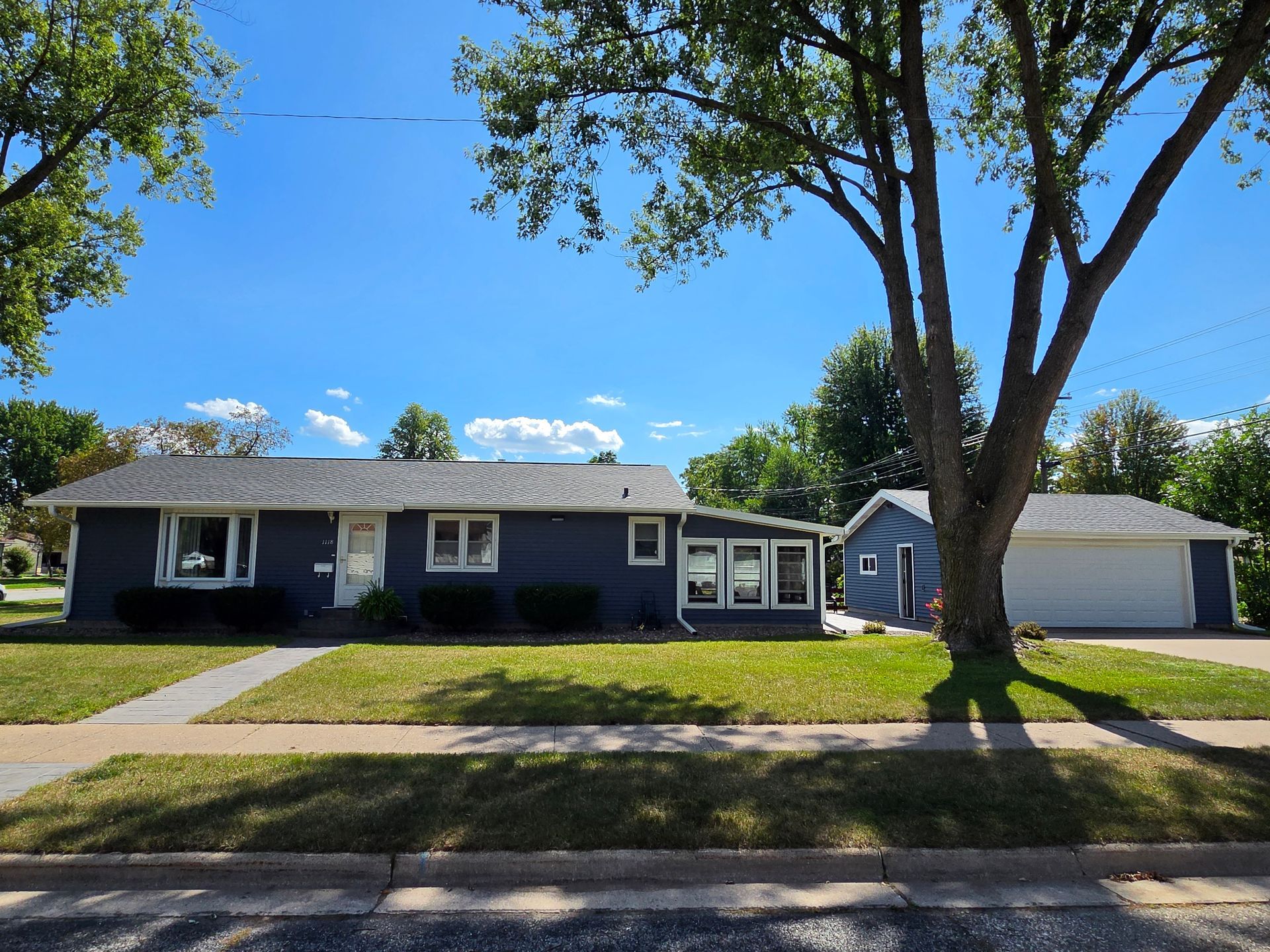 Single-story dark blue house with a large tree, driveway, and detached garage on a sunny day
