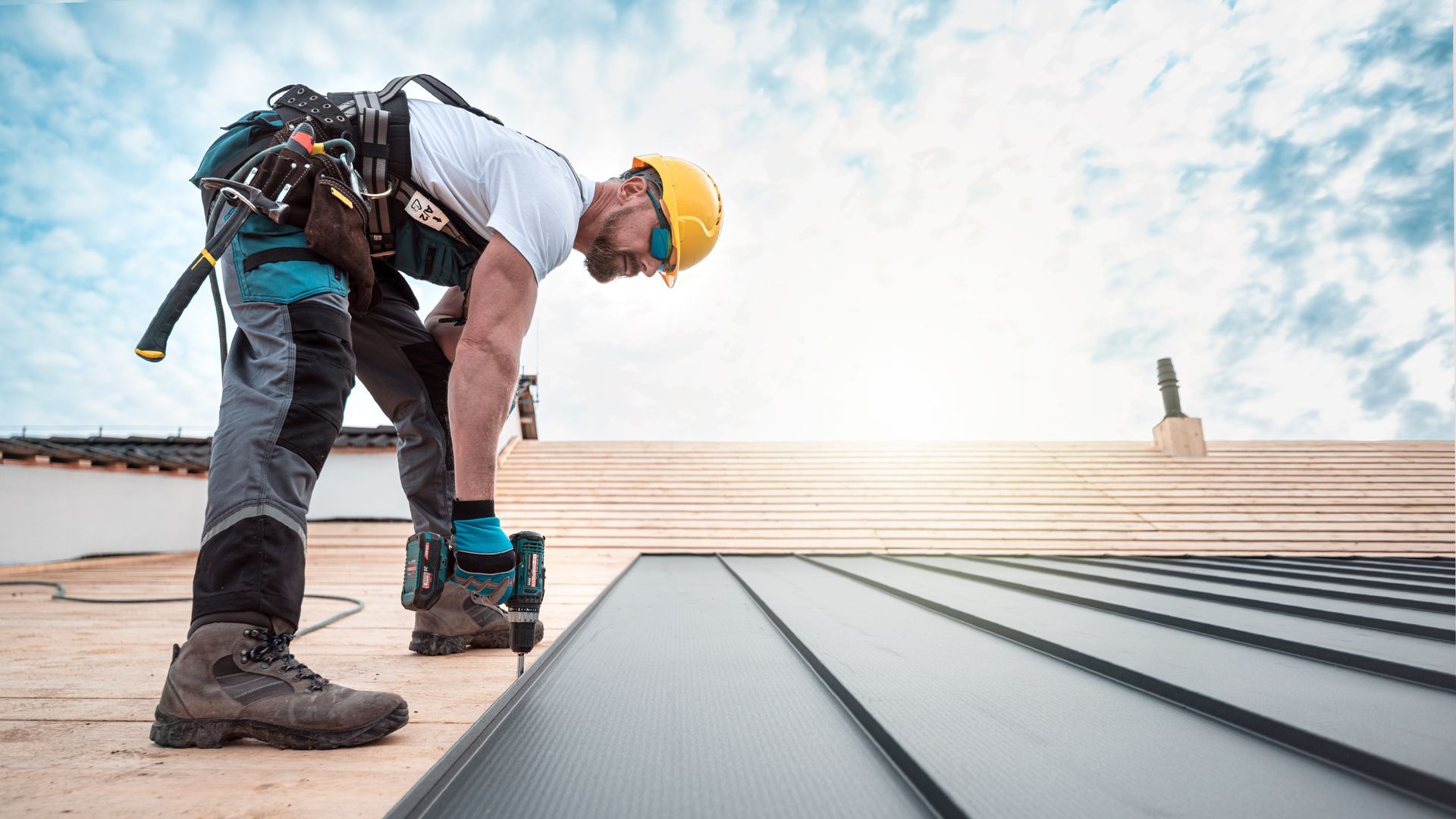 A roofer from a roofing company, on a safety harness and a tool belt, is anchoring a metal roofing.