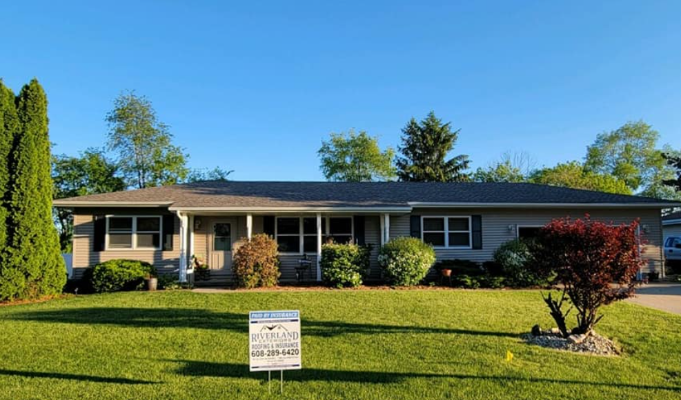 Gray ranch-style house with dark shutters, front porch, and sign on the lawn under a blue sky.