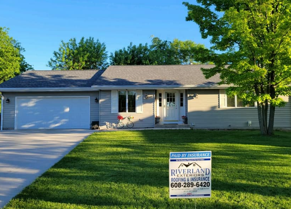 A light gray house with a new roof and a Riverland Roofing sign on the lawn.