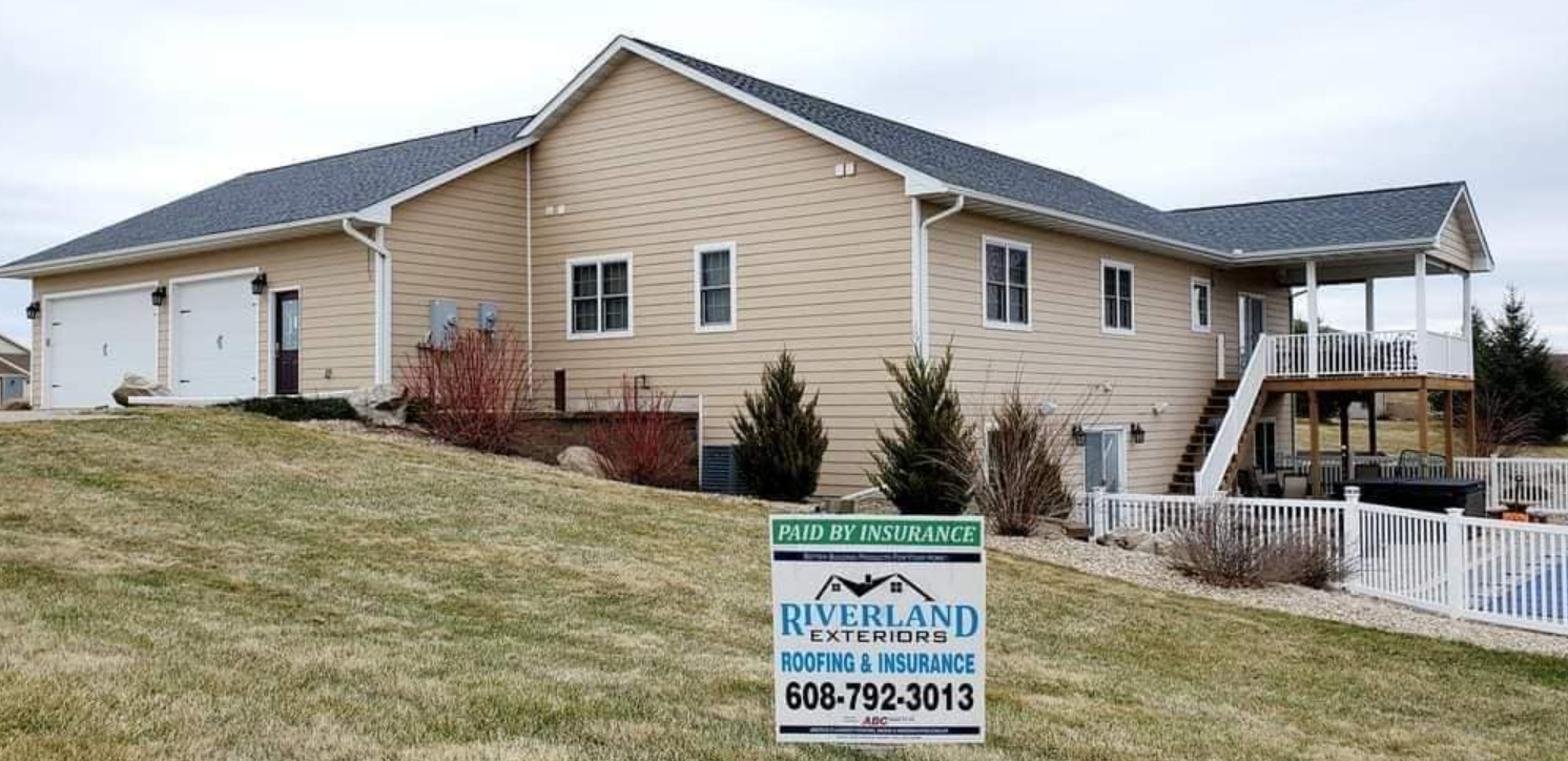 Tan two-story house with a black roof and a sign in the foreground that reads “Riverland.”