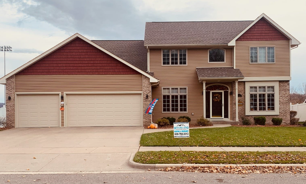 Two-story house with tan siding, brown roof, and garage doors. A front yard has a lawn with fallen leaves.