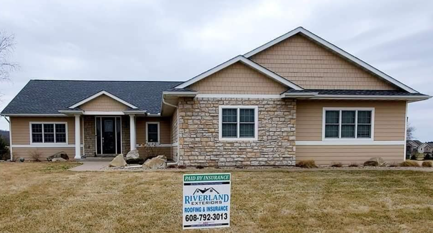 Tan and stone-faced ranch-style house with dark gray roof, set on a grassy lawn with a real estate sign.