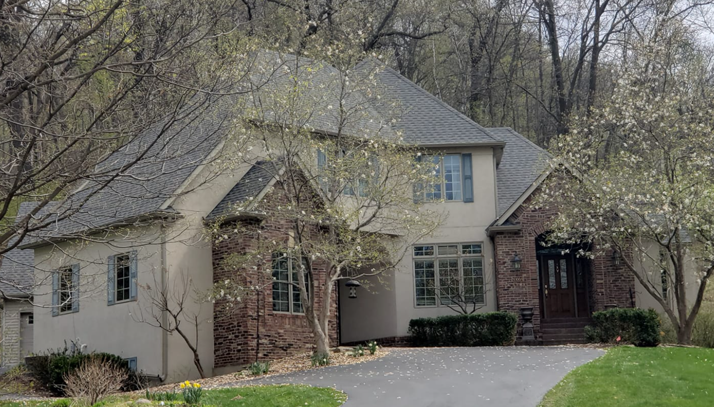 Two-story house with tan stucco, brick accents, and a winding driveway, surrounded by trees.