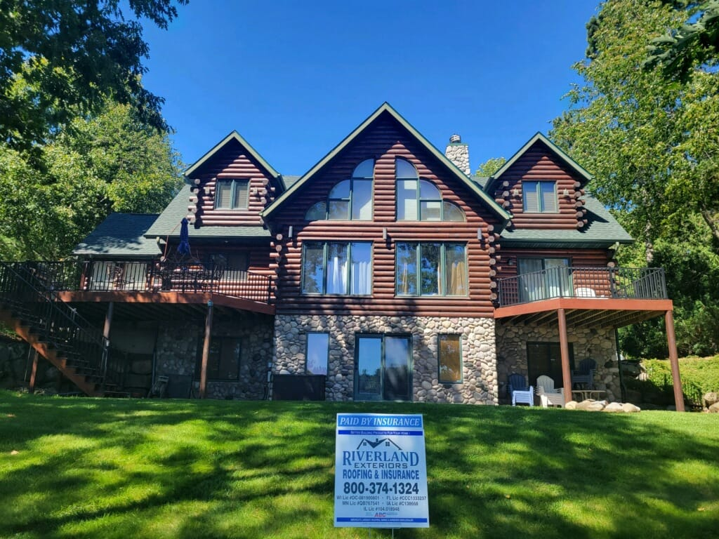 Log cabin house with stone foundation, large deck, and green lawn on sunny day.