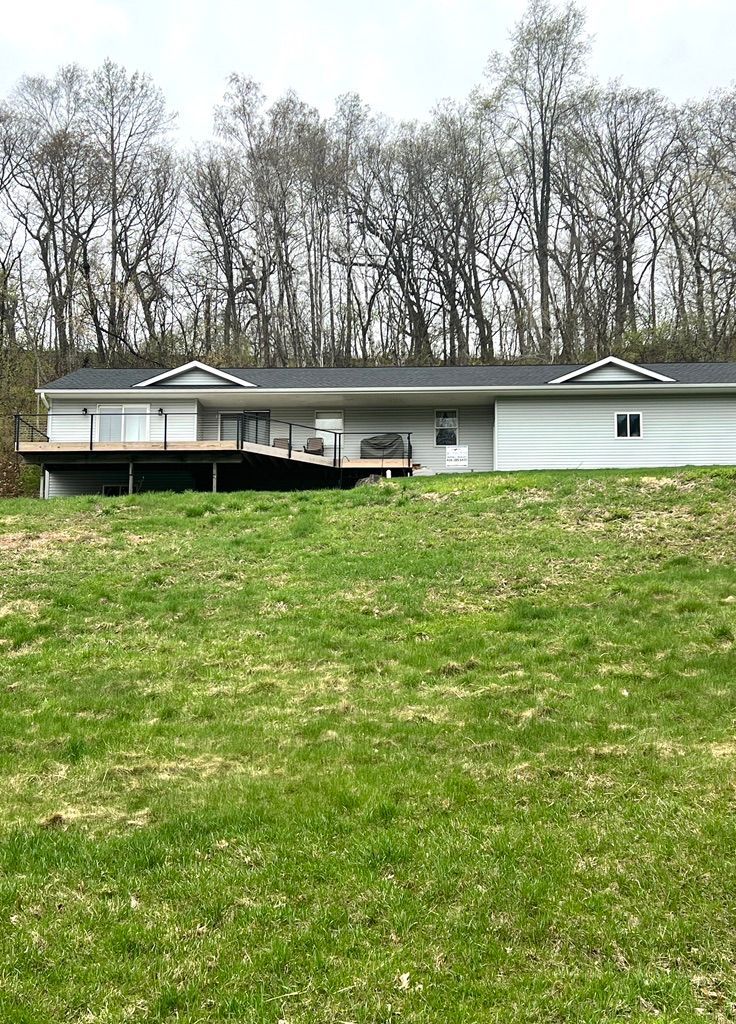 A light gray house with a deck sits on a grassy hill, with trees in the background.