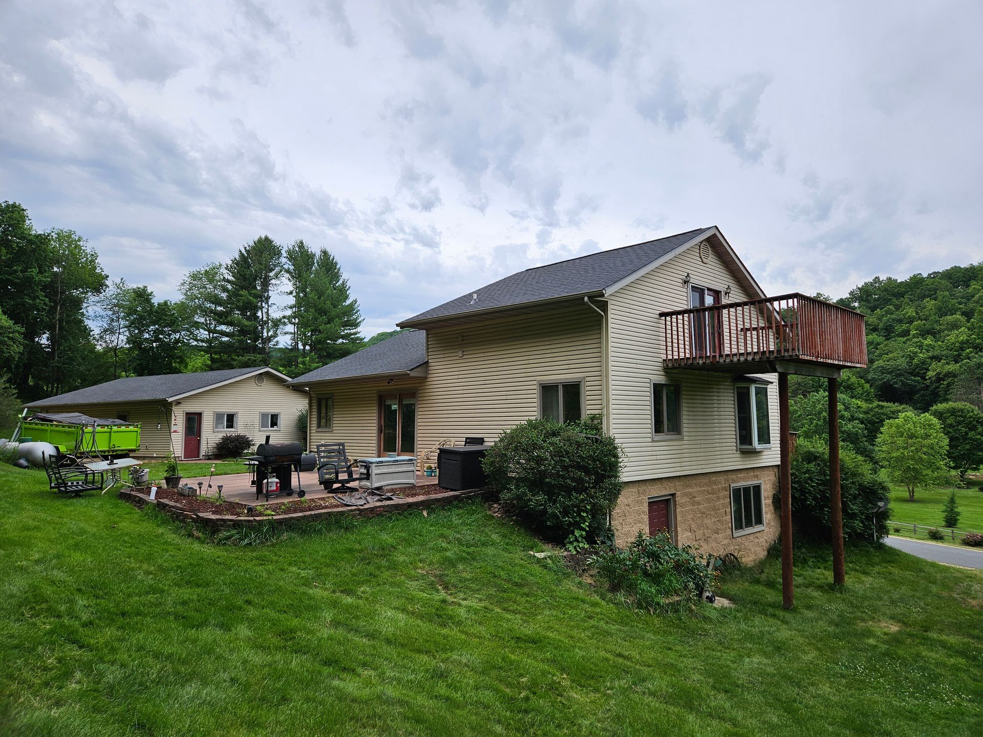 House with deck and patio, set on a grassy hill, with trees in the background under a cloudy sky.