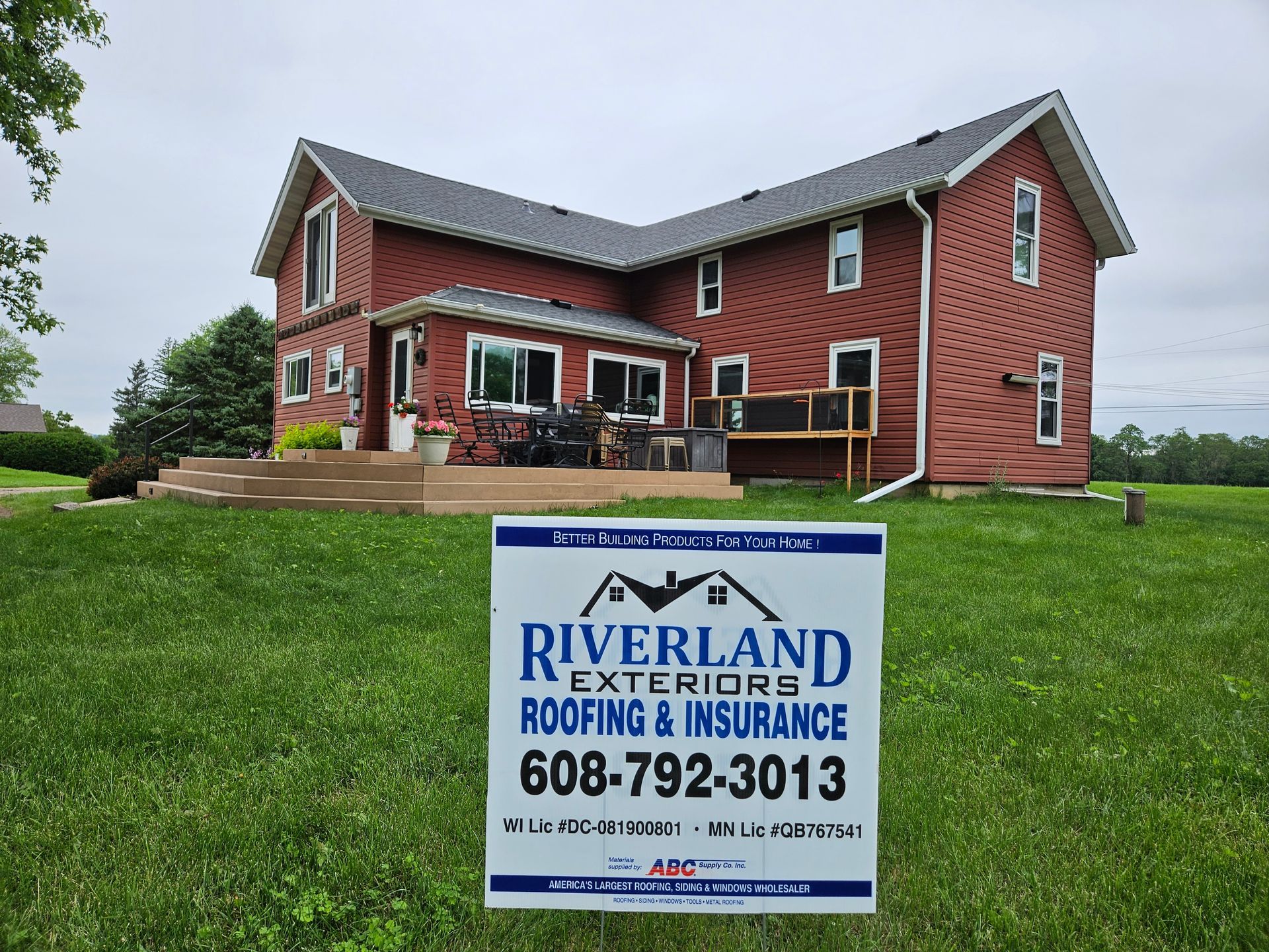 Red two-story house with a new roof, a sign in the foreground advertises Riverland Exteriors Roofing & Insurance.