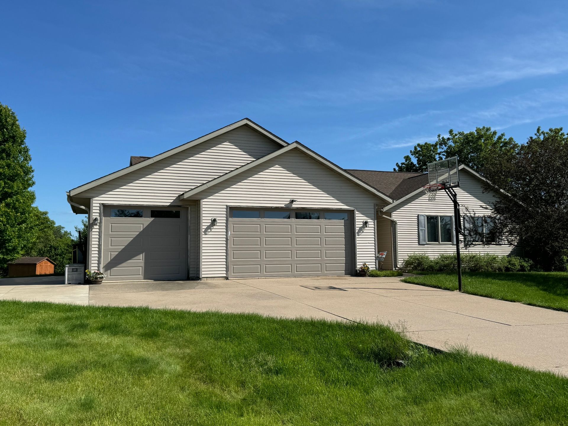 Beige house with two-car garage, driveway, and basketball hoop on a sunny day.