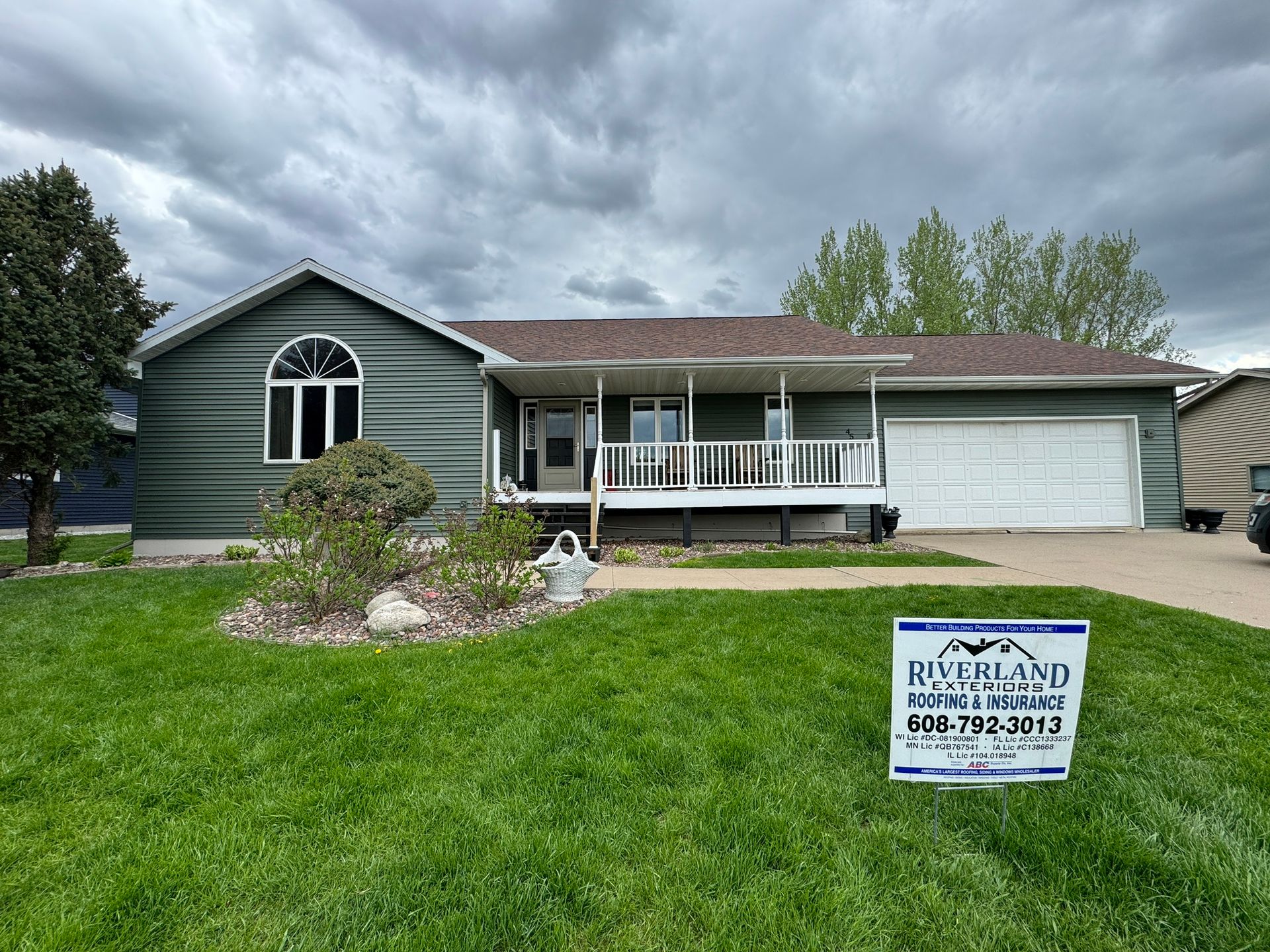 Green house with porch, white garage door, and a sign on the lawn. Overcast sky.