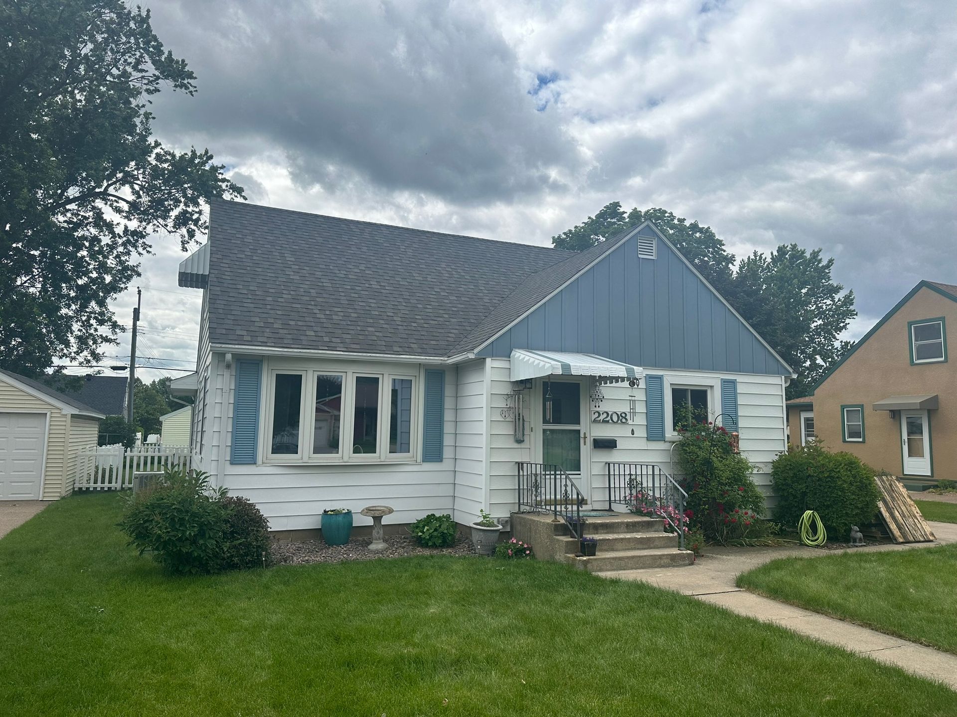 White house with blue trim, small porch, lawn, and gray roof under cloudy sky.