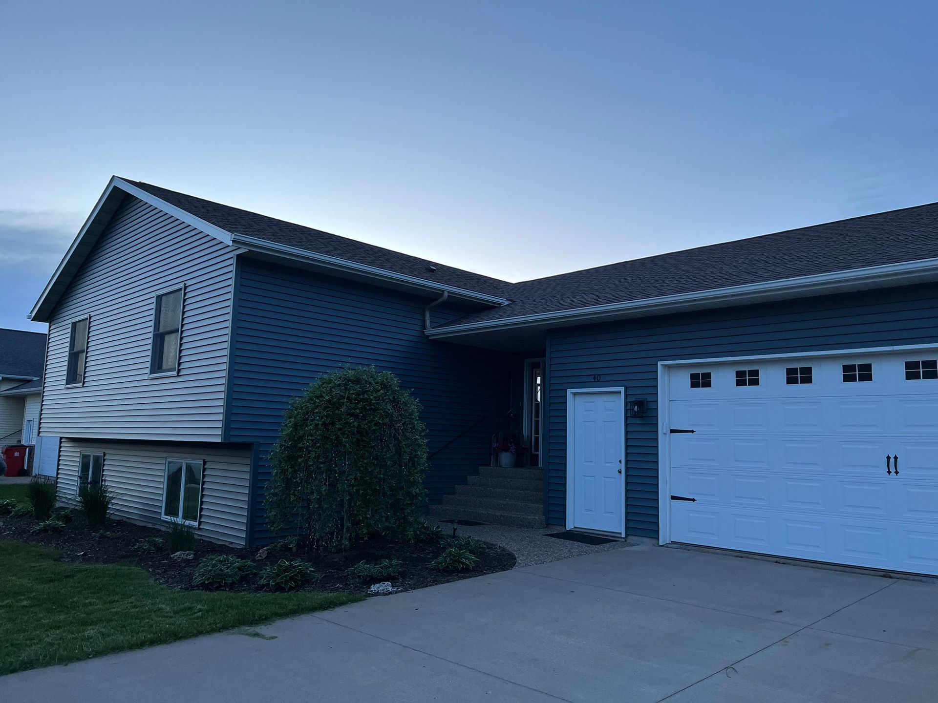 Two-story house with blue siding, white garage door, and a small front yard with shrubs and a driveway.