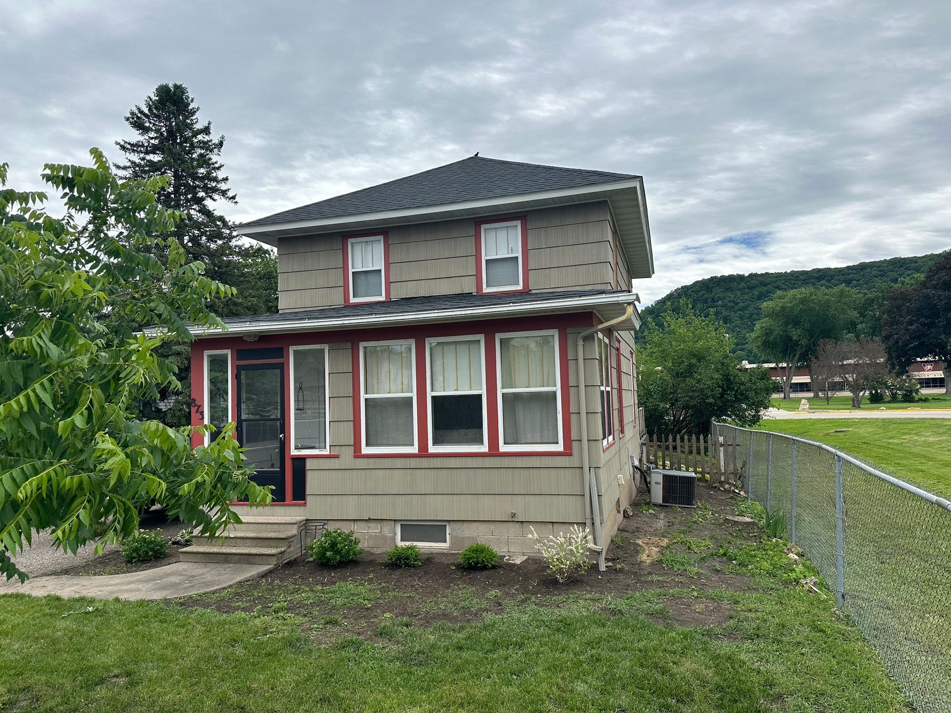 Two-story house with gray siding, red trim, and a small porch, next to a chain-link fence on a grassy lot.