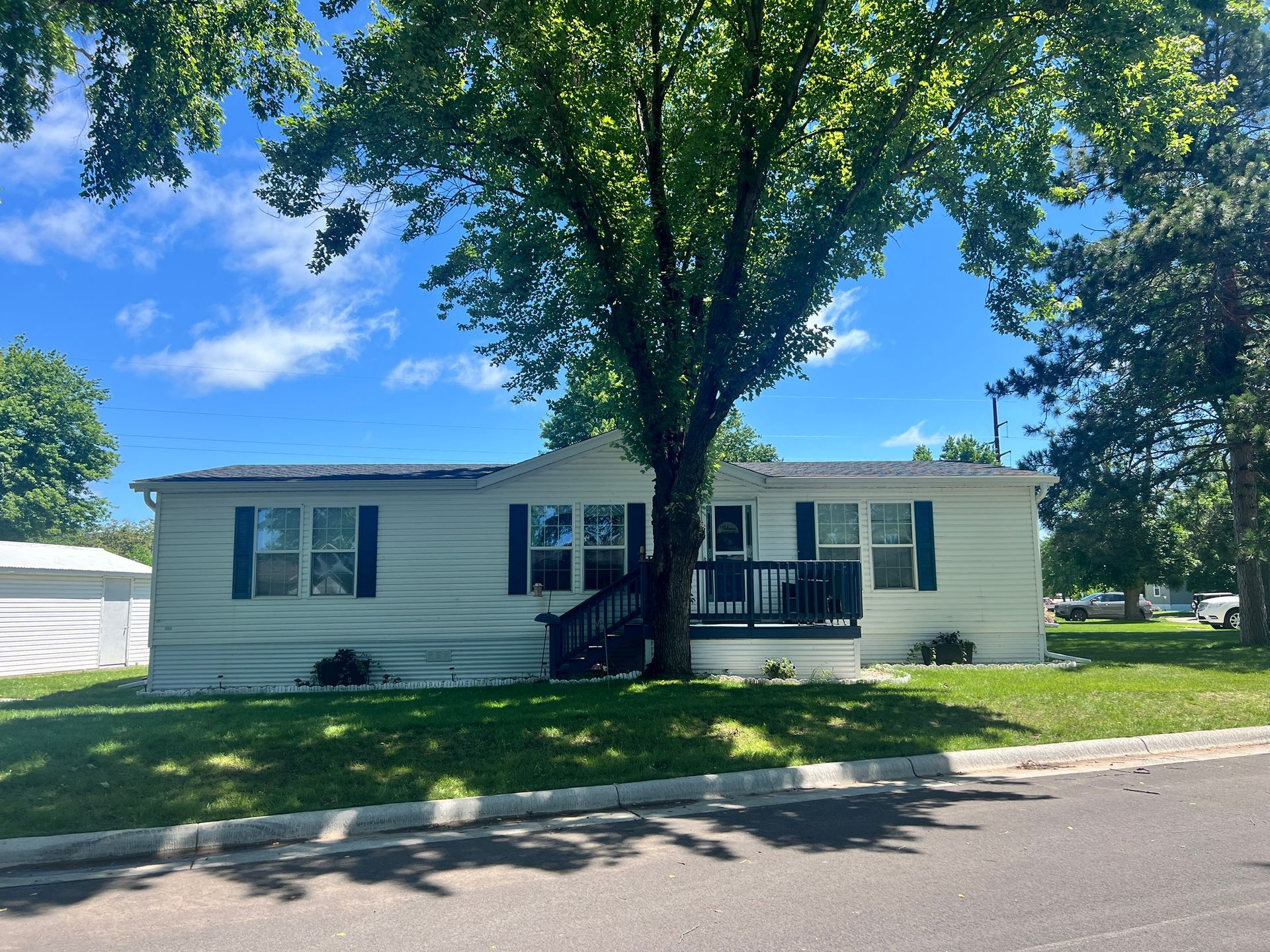 White single-story house with blue shutters and a porch, a large tree in front, on a sunny day.
