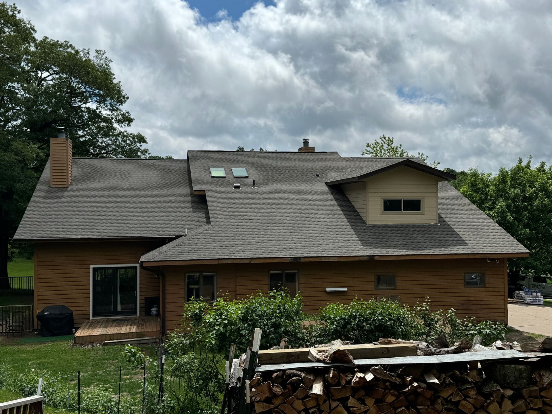 Wooden house with a shingled roof under cloudy skies, surrounded by trees and a garden.