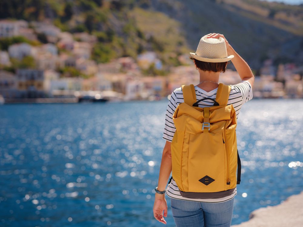 Una persona con una mochila amarilla y un sombrero de paja contempla un puerto azul resplandeciente, con barcos y edificios en la ladera.