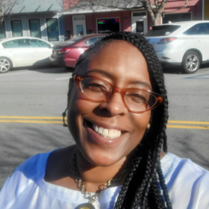 A woman wearing glasses and a necklace is smiling in front of a mural.